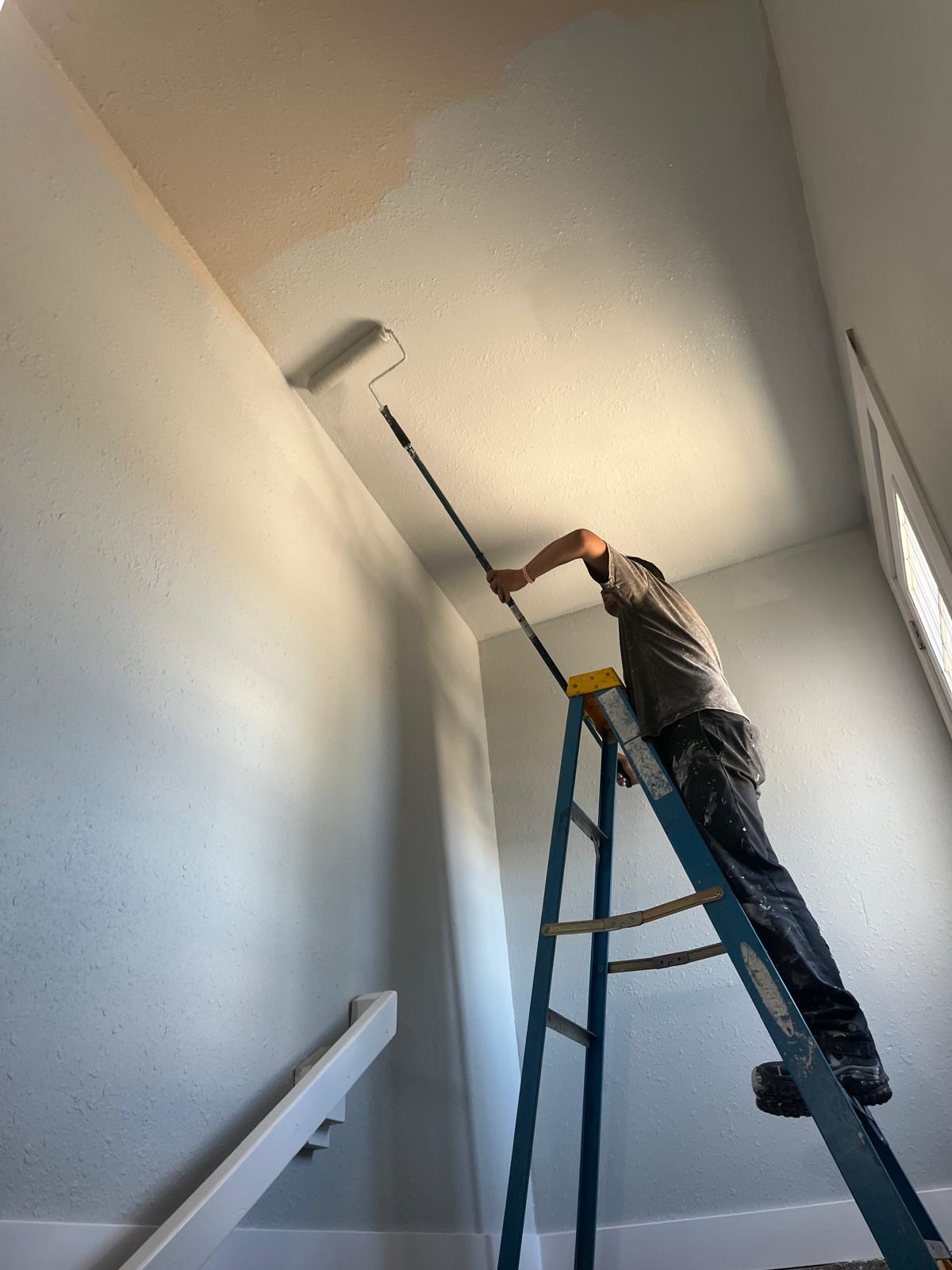 Crew member rolling fresh paint on a tall interior stairwell wall from a ladder.