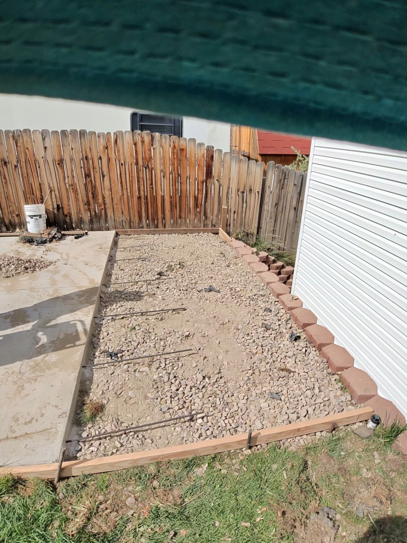 Formed and rebar-tied gravel base prepared for a concrete pad beside an existing slab in a fenced backyard.