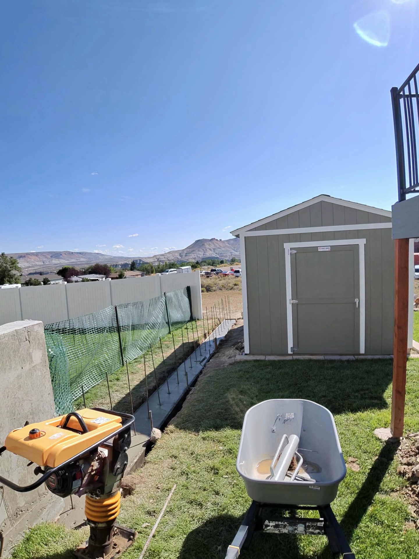 Formed and rebar-pinned footing along a backyard fence line with a plate compactor staged nearby.