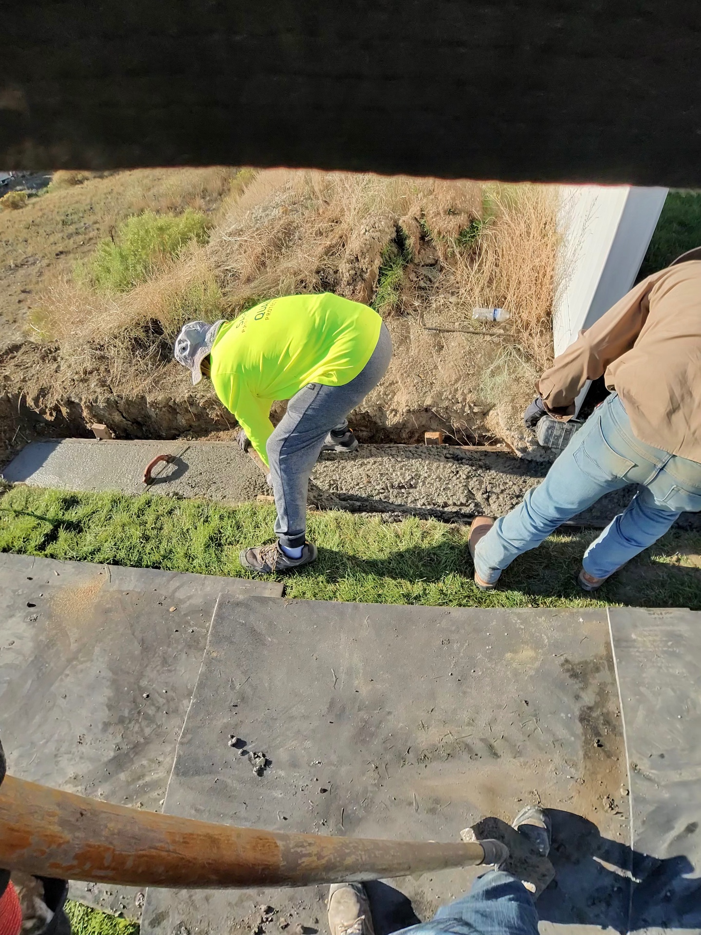 Crew placing and screeding fresh concrete in a narrow curb and walkway form beside a building.
