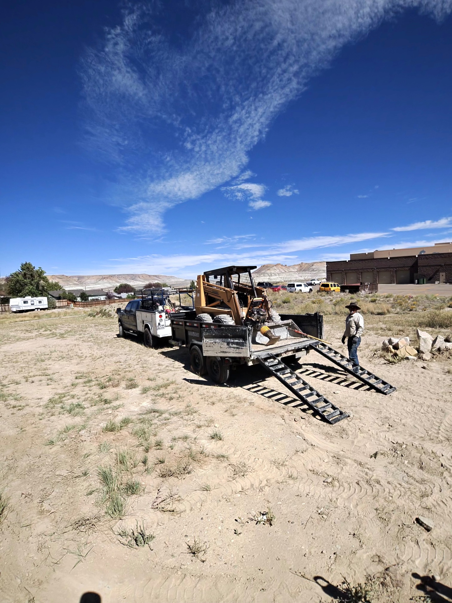 Skid steer loaded on a dump trailer behind a work truck on an open Wyoming lot.