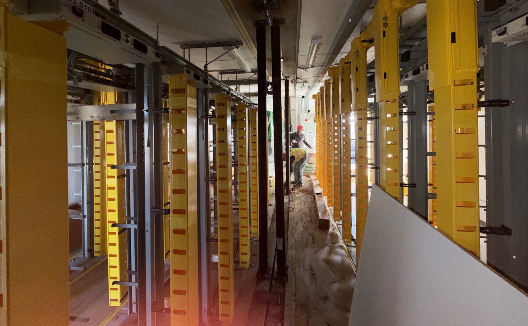 Crew member performing construction support inside a telecommunications facility aisle; equipment racks shown were installed by another trade.