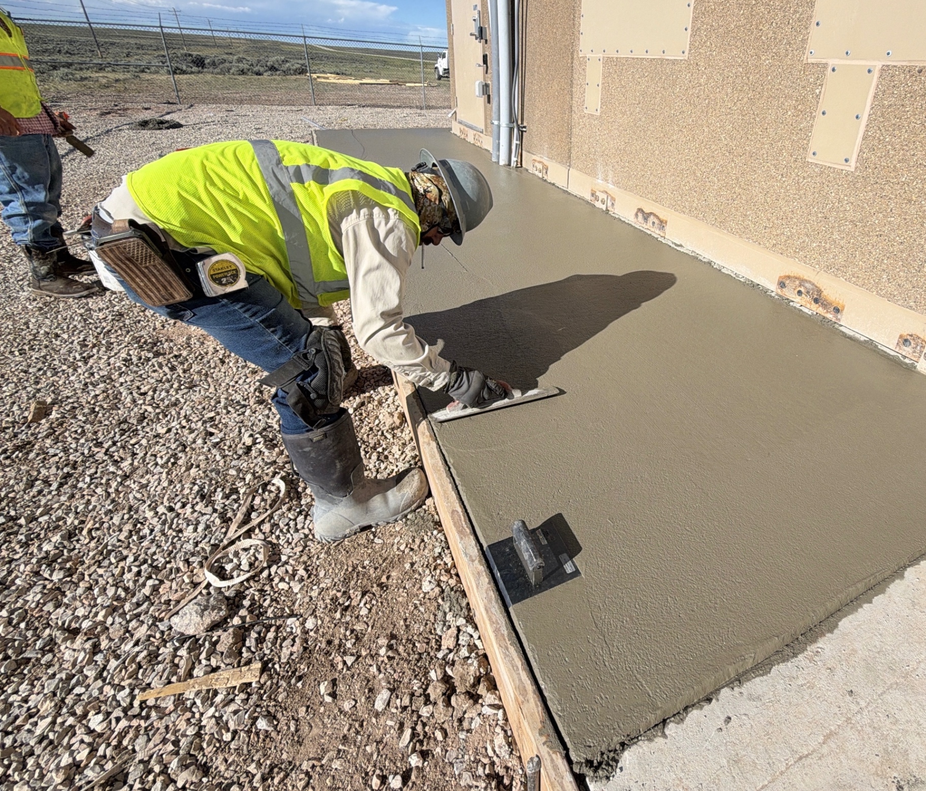 JJJ crew member wearing high-visibility gear and a hard hat hand-finishing fresh concrete beside a commercial facility.